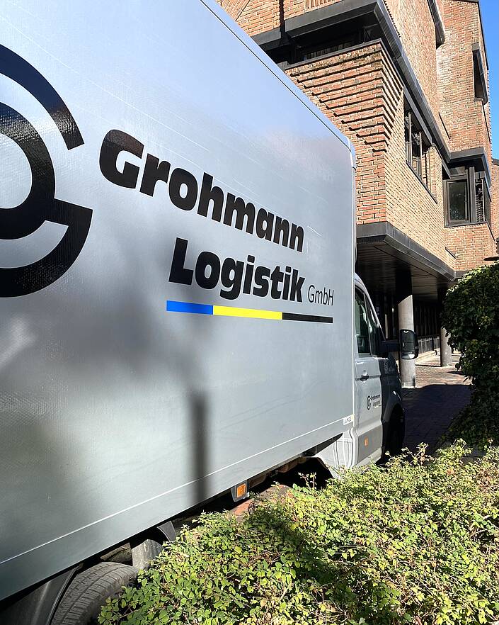 Im Vordergrund eine Seite eines LKW mit Logoaufschrift vor Gebäude